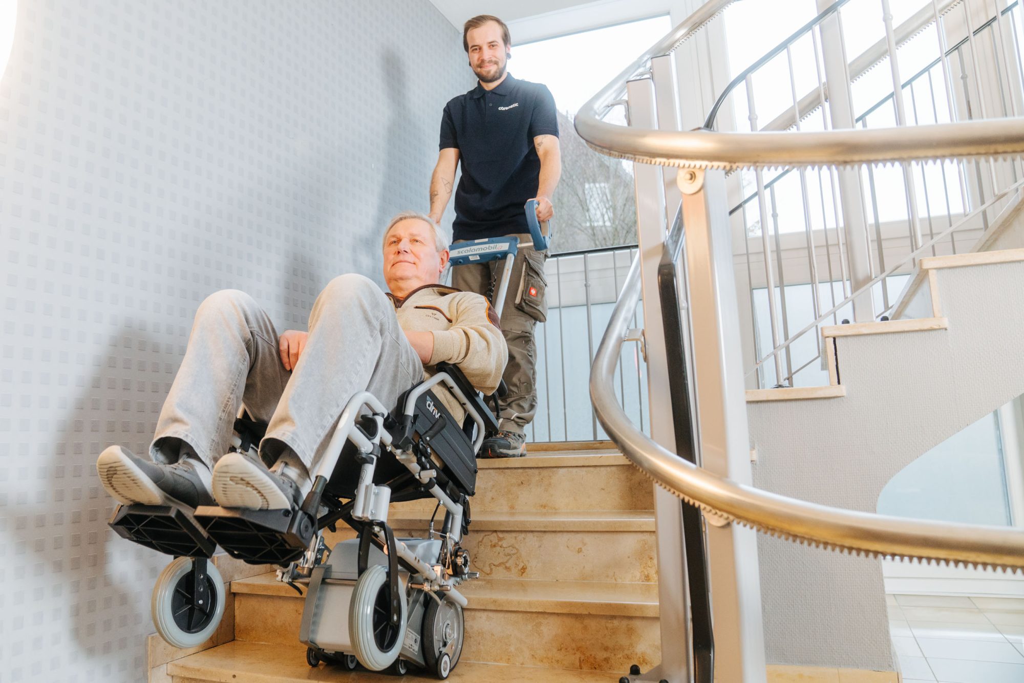Carenetic Mitarbeiter befördert einen Mann im Rollstuhl mit einem Treppensteiger über eine Treppe