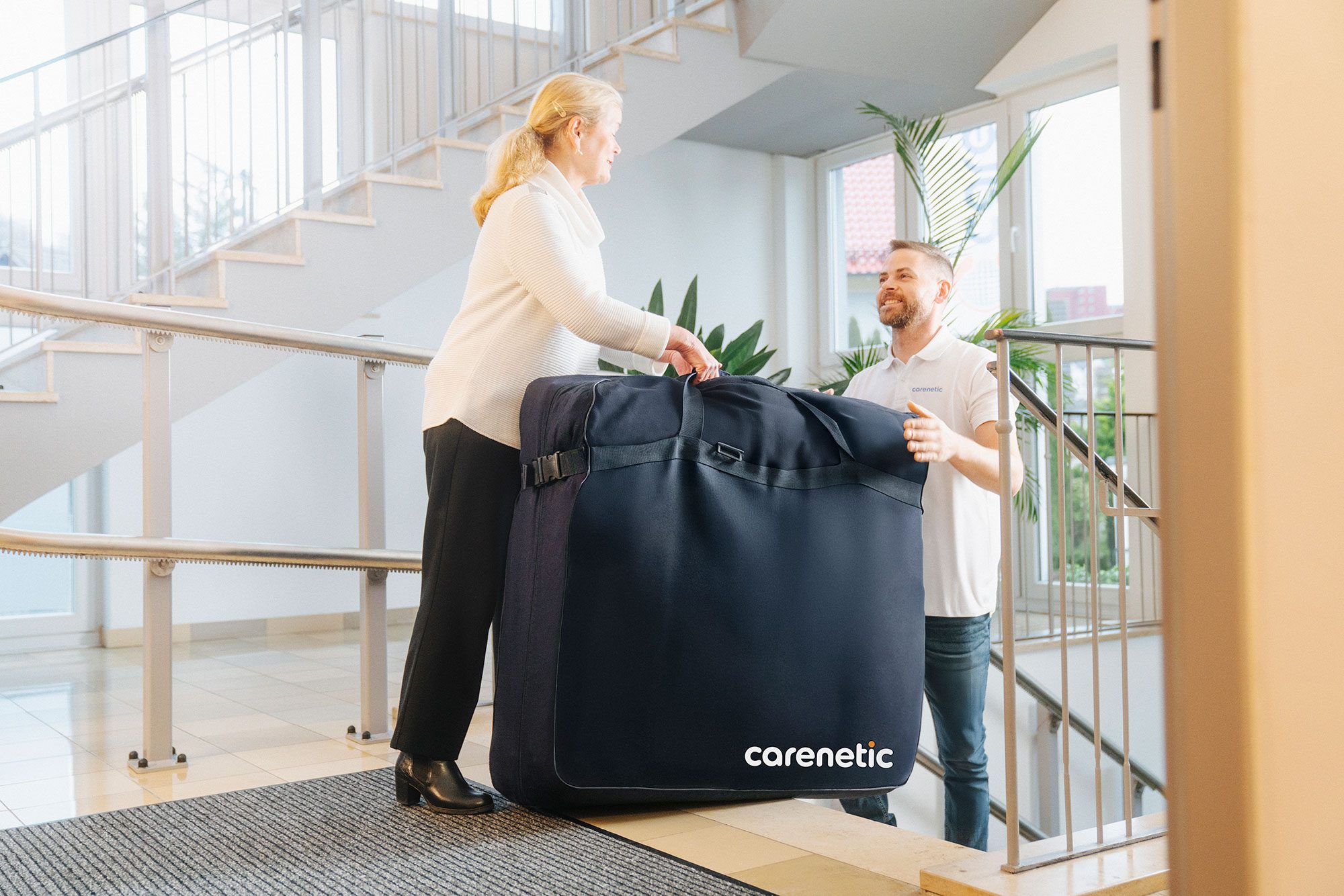 Carenetic Mitarbeiter und eine Kundin tragen eine Matratze in der Carenetic Transporttasche durchs Treppenhaus
